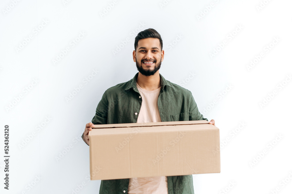 Positive attractive candid arabian or indian young delivery man in casual clothes, holding a cardboard box, standing on isolated white background, looks at camera, smiles friendly. Delivery concept Positive attractive candid arabian or indian young delivery man in casual clothes, holding a cardboard box, standing on isolated white background, looks at camera, smiles friendly. Delivery concept