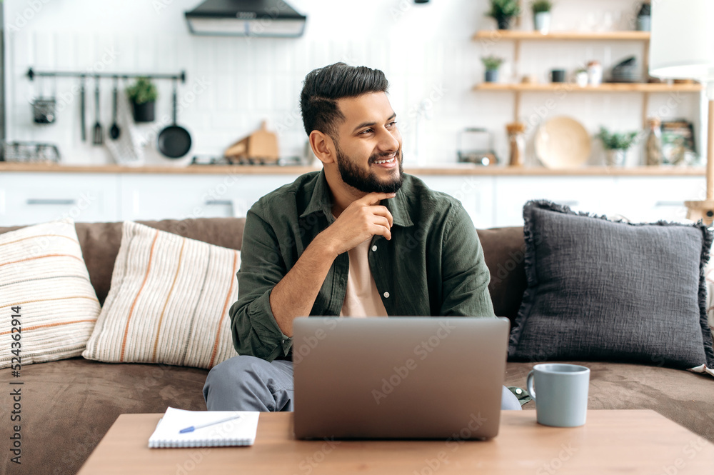 Handsome confident thoughtful arabian or indian guy in casual wear, freelancer, sit on sofa at home in living room, using a laptop for work, looking pensively away, dreaming, thinking, smiling Handsome confident thoughtful arabian or indian guy in casual wear, freelancer, sit on sofa at home in living room, using a laptop for work, looking pensively away, dreaming, thinking, smiling