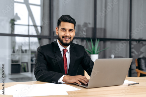 Portrait of attractive elegant confident arabian or indian successful businessman, entrepreneur, lawyer, in formal suit, sit at work desk with laptop in modern creative office, looks at camera, smile