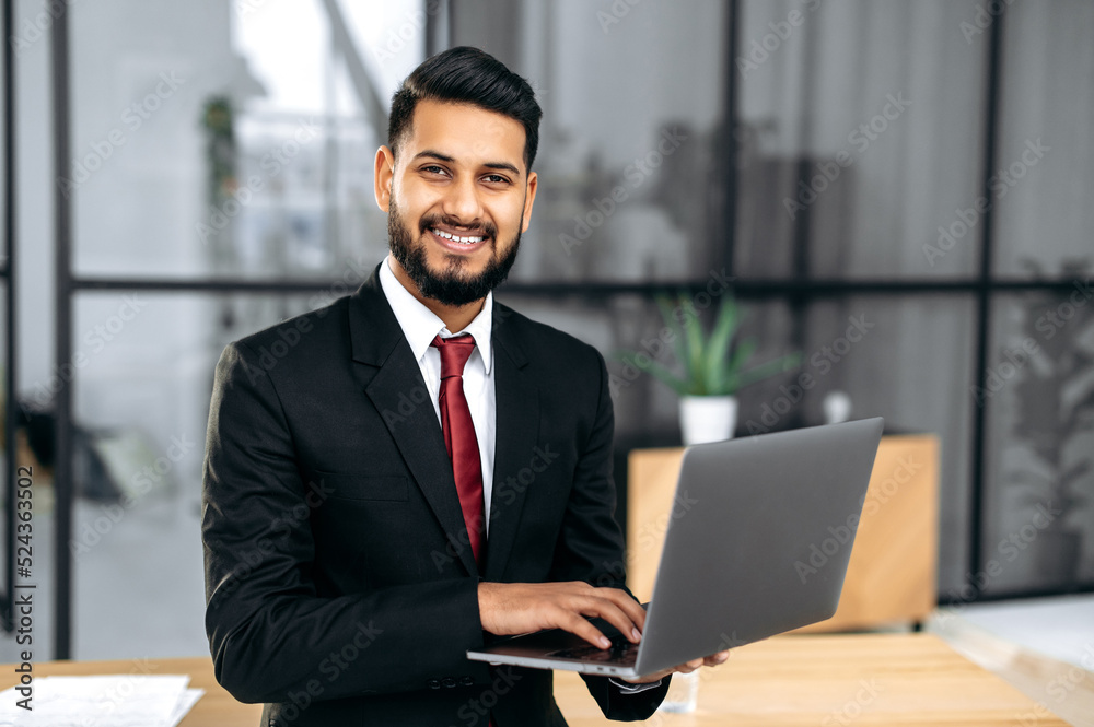 Portrait of successful confident elegant Indian or Arabian young businessman, in a suit, holding an open laptop in his hand, stand near the desktop in his modern office, looking at the camera, smiling Portrait of successful confident elegant Indian or Arabian young businessman, in a suit, holding an open laptop in his hand, stand near the desktop in his modern office, looking at the camera, smiling