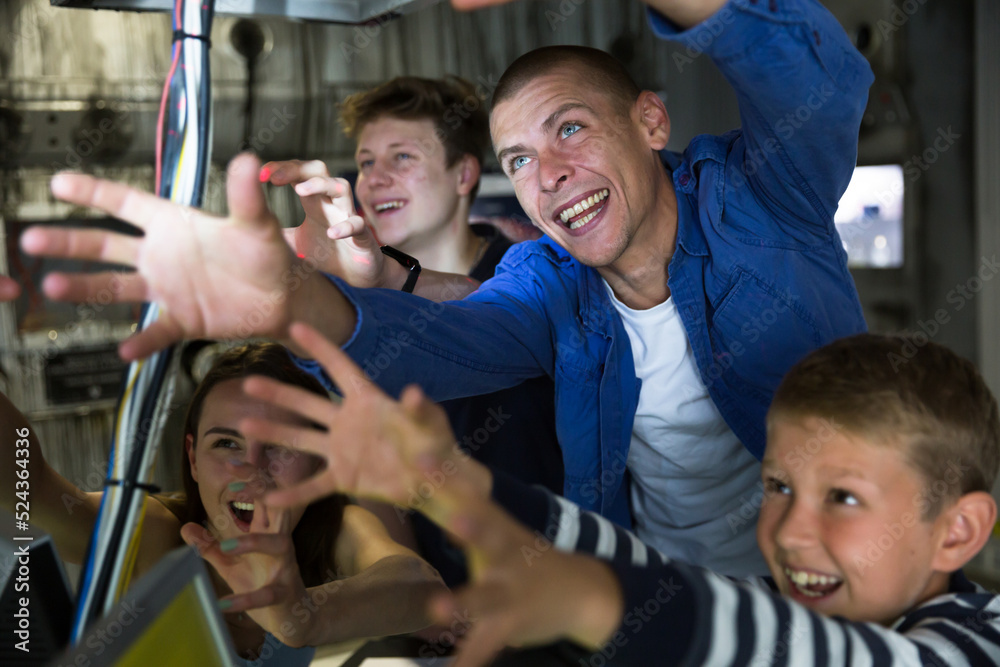Fototapeta premium Portrait of cheerful family of four with preteen sons reaching for something in quest room stylized as underground shelter, holding out their hands..