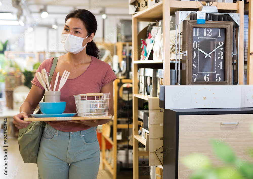 Asian woman in face mask walking through hall of housewares store and holding tray with tableware on it in hands.