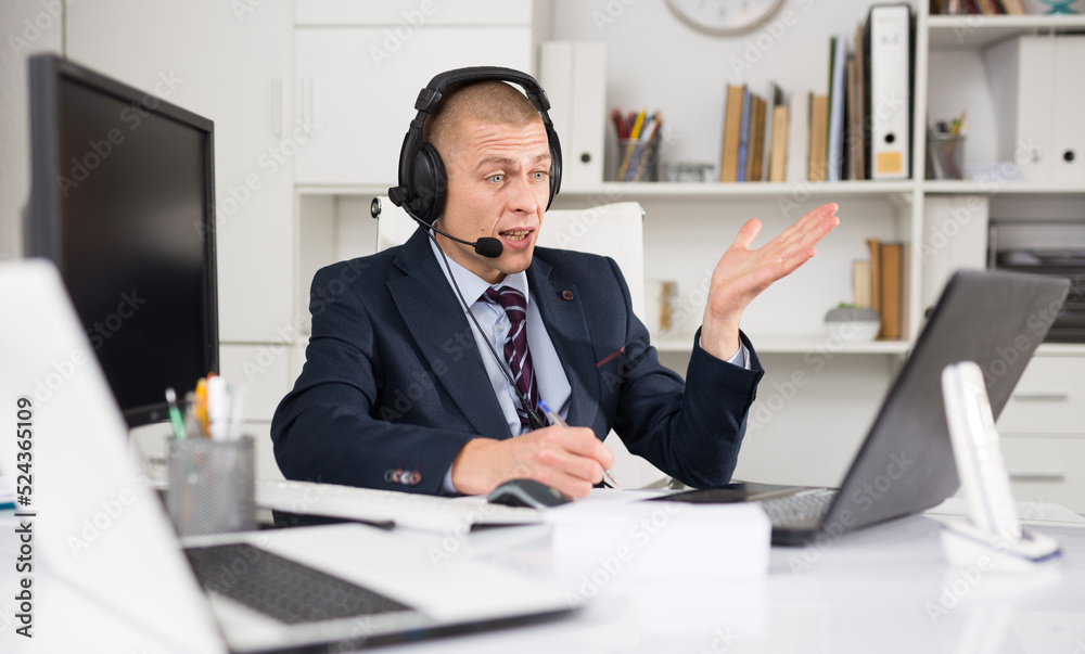 Positive businessman with headphones and microphone having video call on laptop in modern office