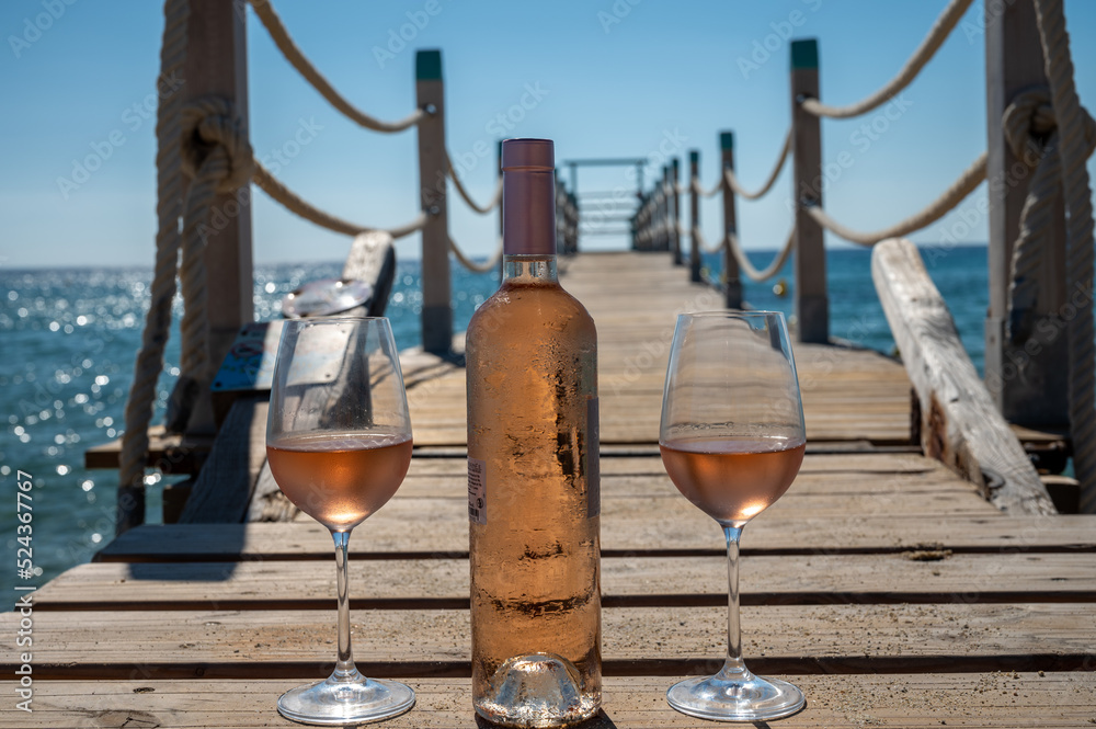 Obraz premium Glasses and bottle of cold rose wine from Provence served outdoor on wooden yacht pier with view on blue water and white sandy beach Plage de Pampelonne near Saint-Tropez, France