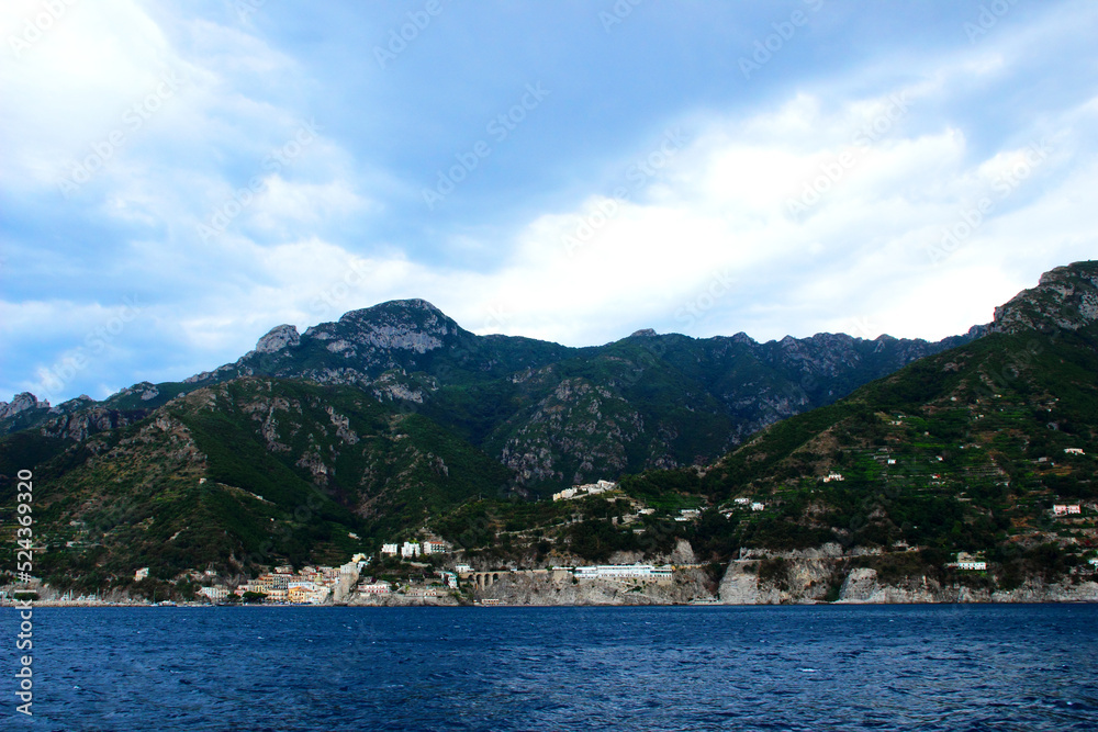 Panorama in Amalfi coast with rough mountain cliffs overlooking a host of peculiar white buildings sitting on rocky shores which skim a deep rippling sea under an unreal sky of fluffy white clouds