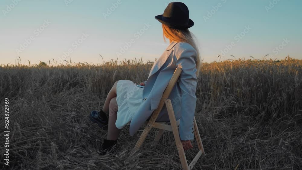 A girl in a hat and jacket is sitting on a chair at sunset in a field. Cool smooth camera movement