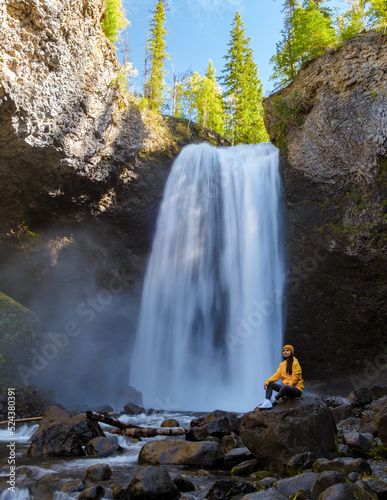 Moul waterfall in Canada, couple visit Moul Falls, the most famous waterfall in Wells Gray Provincial Park in British Columbia, Canada. Asian woman with a yellow sweater and hat looking at a waterfall photo
