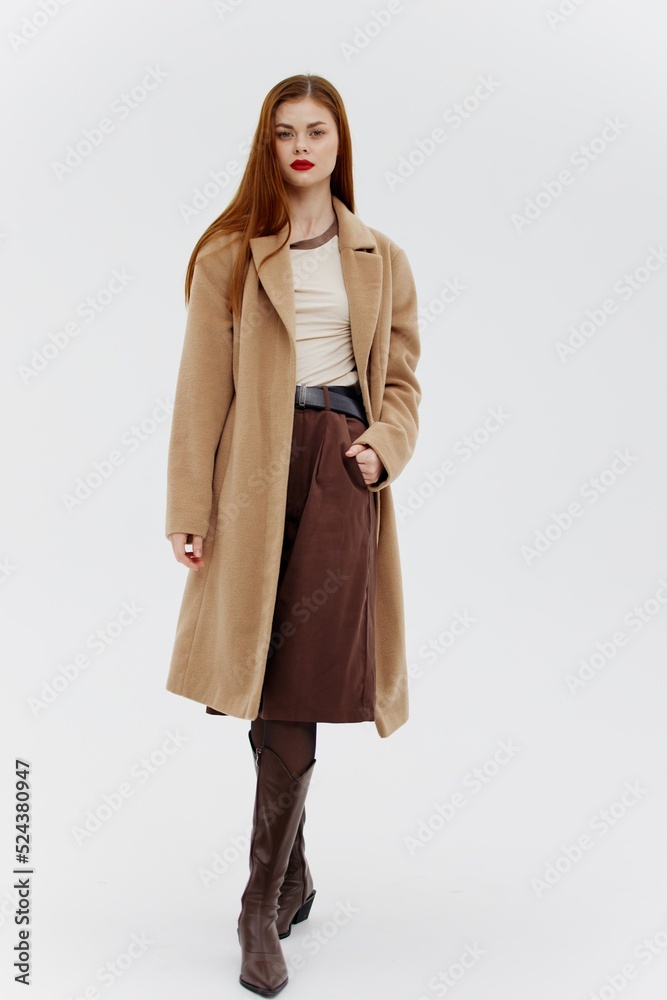 Full-height shooting. A red-haired girl dressed in brown clothes in a coat and boots poses on a white background in the studio Full-height shooting. A red-haired girl dressed in brown clothes in a coat and boots poses on a white background in the studio