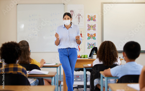 Young female teaching after covid pandemic, in classroom with young children students, in face mask. Back to school for little kids learning their education from their teacher after quarantine