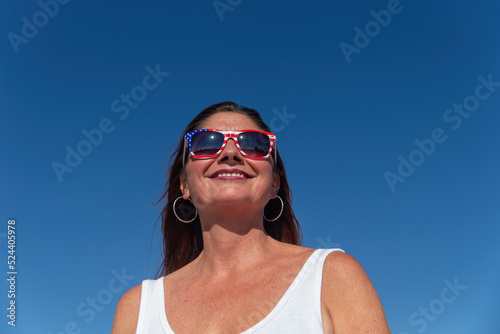 Beautiful smiling middle aged 40+ 50+ year old woman wearing patriotic American flag sunglasses with plain blue sky background