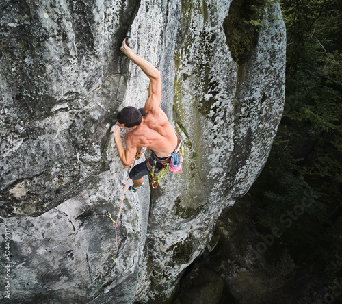 Young man climber climbing difficult route on high boulder with rope. Fearless rock climber training rock climbing on summer day. Concept of extreme sport, adventures and active lifestyle. Aerial view