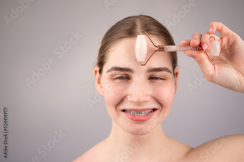 Close-up portrait of a woman with braces on her teeth uses a quartz roller massager to smooth wrinkles on her forehead. 