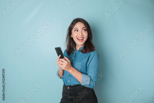 A portrait of a happy Asian woman is smiling and holding her smartphone wearing a blue shirt isolated by a blue background