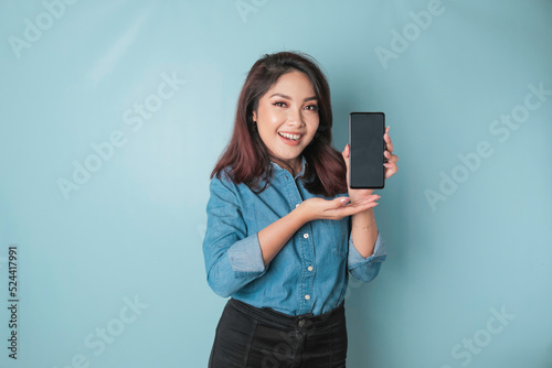 Excited Asian woman wearing blue shirt pointing at the copy space on her smartphone, isolated by blue background