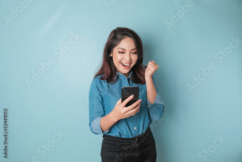 A young Asian woman with a happy successful expression wearing a blue shirt holding smartphone isolated by blue background