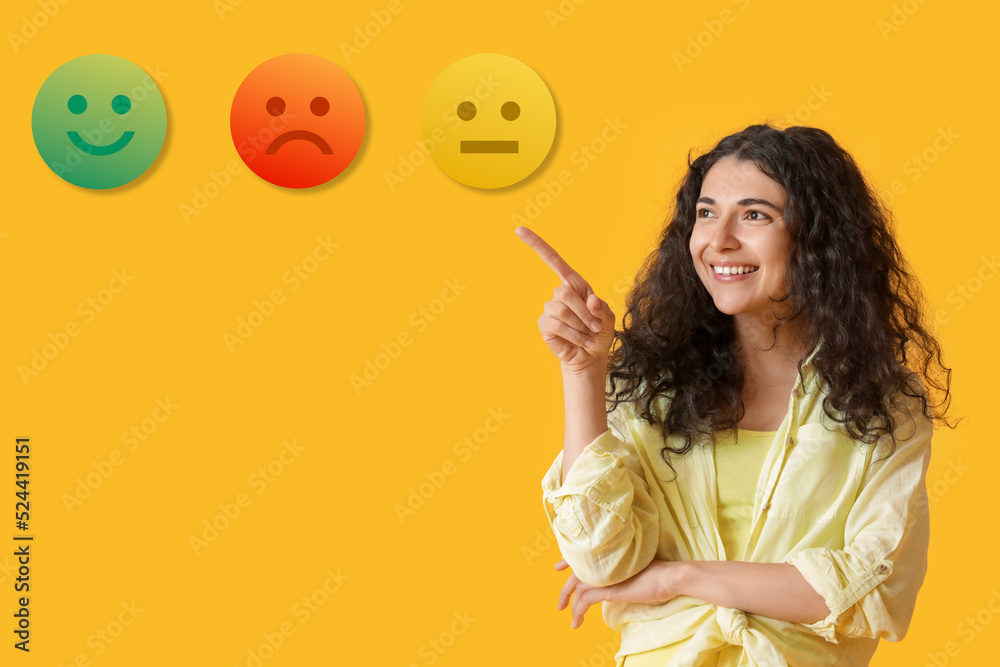 Fototapeta premium Beautiful Asian woman pointing at different emoticons on yellow background