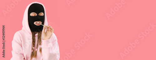Fotografiet Young woman in balaclava and hoodie chewing gum on pink background with space fo