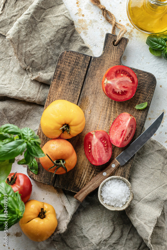 Fototapeta Naklejka Na Ścianę i Meble -  Raw tomatoes and knife on wooden cutting board surrounded by ingredients salt basil leaves and olive oil around. Top view. Rustic cooking background