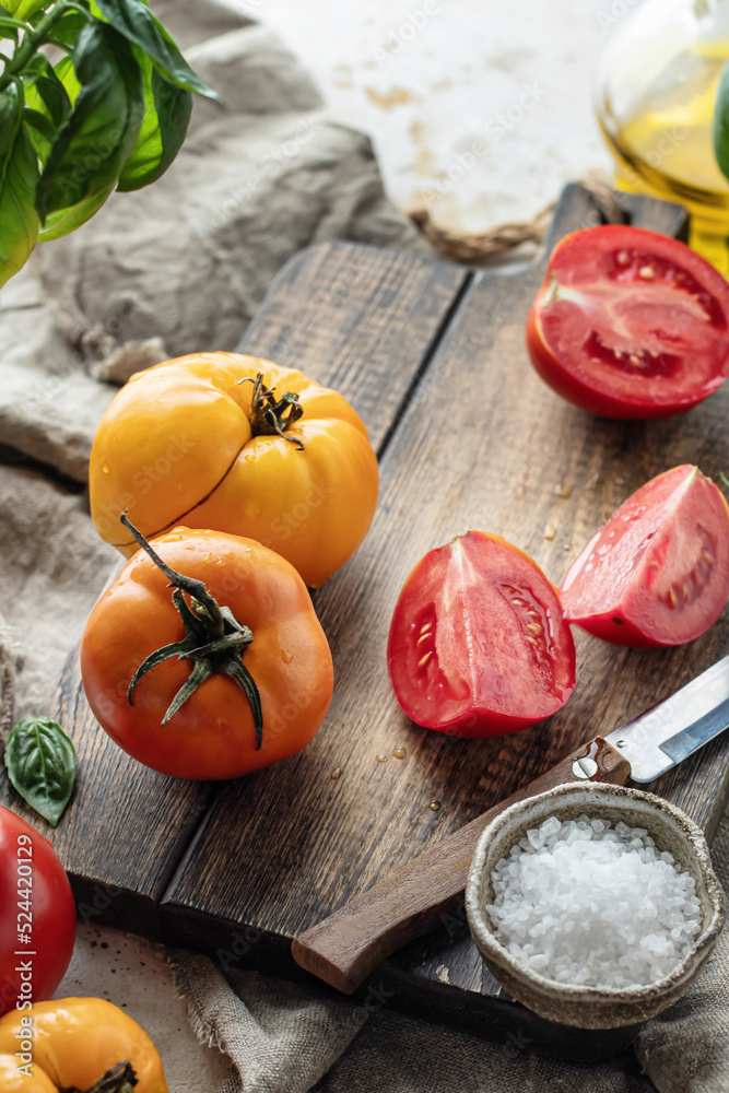 Raw tomatoes and knife on wooden cutting board surrounded by ingredients salt basil leaves and olive oil around. Rustic cooking background