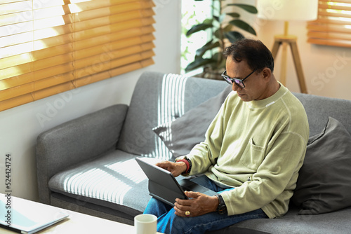 Mature man wearing eyeglasses reading online news on digital tablet while relaxing on couch in bright living room