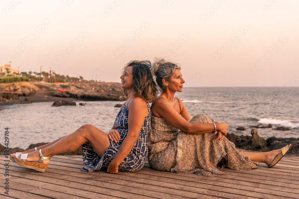 two lovely middle aged girlfriends sitting on a wooden bridge by the sea at sunset