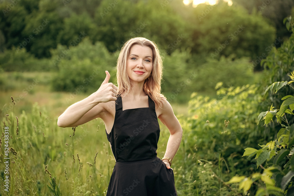 a happy blonde woman in a black dress against the background of nature, in the park shows a sign with a thumbs-up gesture, everything is OK, and smiles a happy blonde woman in a black dress against the background of nature, in the park shows a sign with a thumbs-up gesture, everything is OK, and smiles