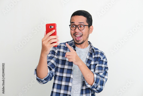 Young Asian man looking and pointing his mobile phone with surprised expression