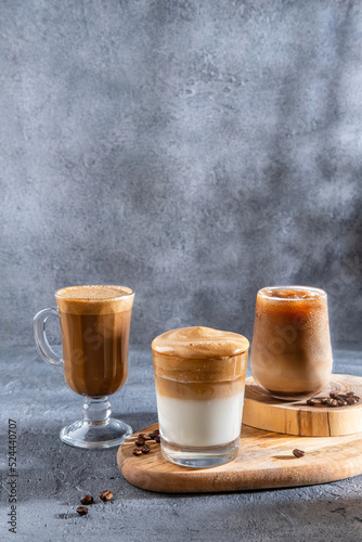 Ice coffee in a tall glass with cream poured over and coffee beans. Set with different types of coffee drinks on a dark table.