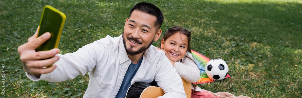 Asian dad taking selfie on cellphone while holding acoustic guitar near child in park, banner. Asian dad taking selfie on cellphone while holding acoustic guitar near child in park, banner.