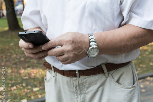 A mobile phone in the large hands of an old man with a watch on his left hand, in a white shirt, on a walk in the park