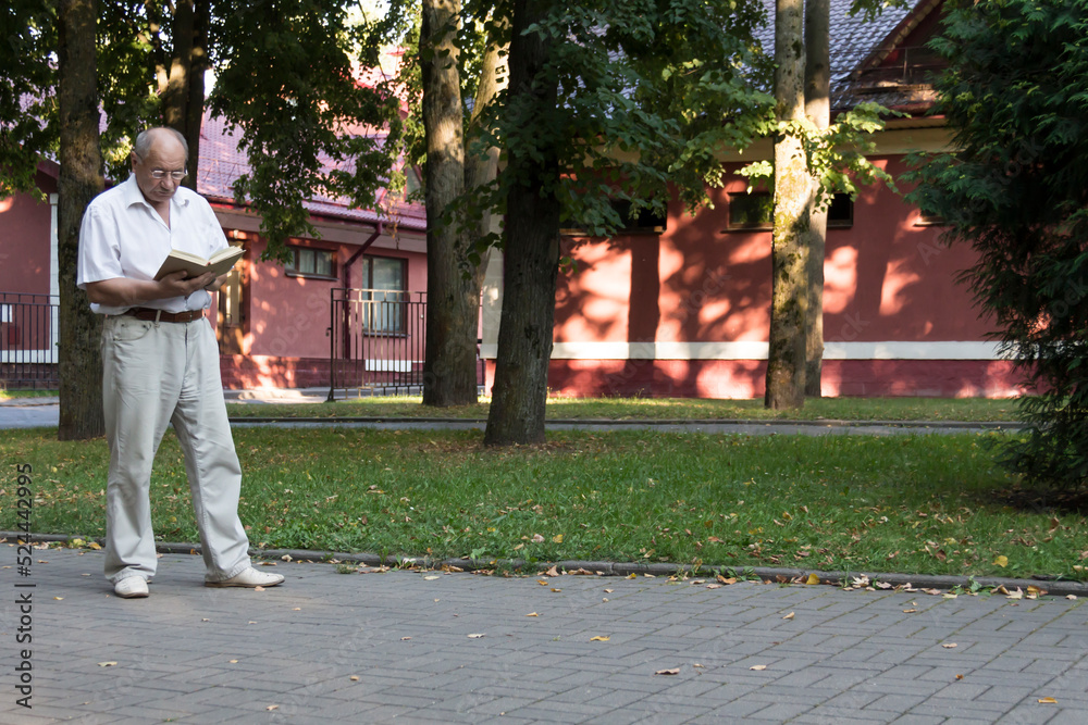 An elderly retired man walks alone in the park. An old, balding man with glasses stands against the background of a red building and reads a book