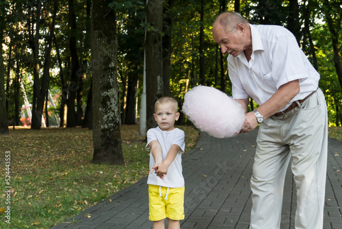 Grandfather lovingly and carefully treats and feeds his little grandson with pink cotton candy in an amusement park. A pensioner and a little boy spend their free time on weekends. #524443553