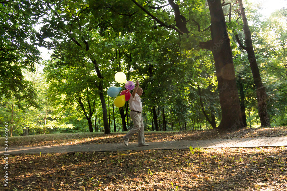A lonely pensioner walks along the path in the park with balloons. An elderly man is having fun in the park among the trees.