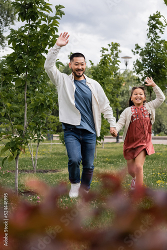Happy asian father and child waving hands while running in park.
