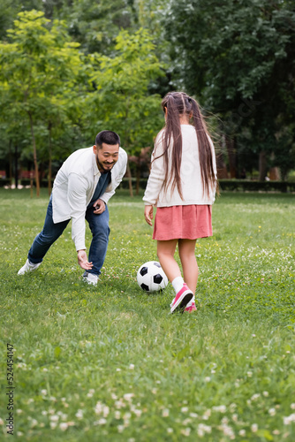 Happy asian father playing soccer with daughter on grass in park.