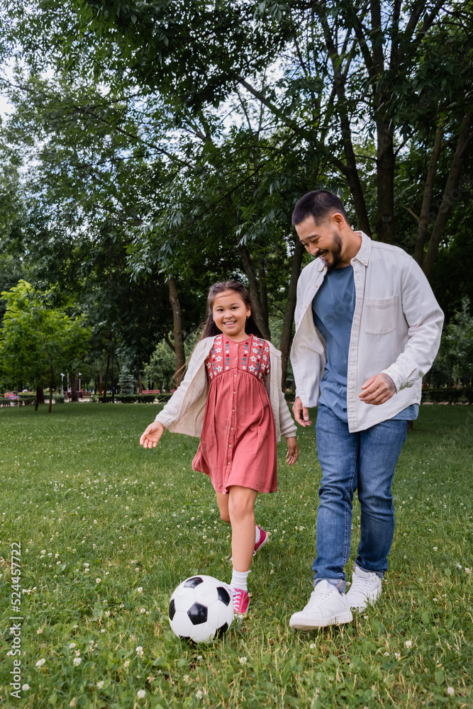 Smiling asian parent and girl playing football on field in park. Smiling asian parent and girl playing football on field in park.