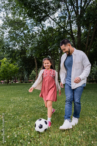 Smiling asian parent and girl playing football on field in park.