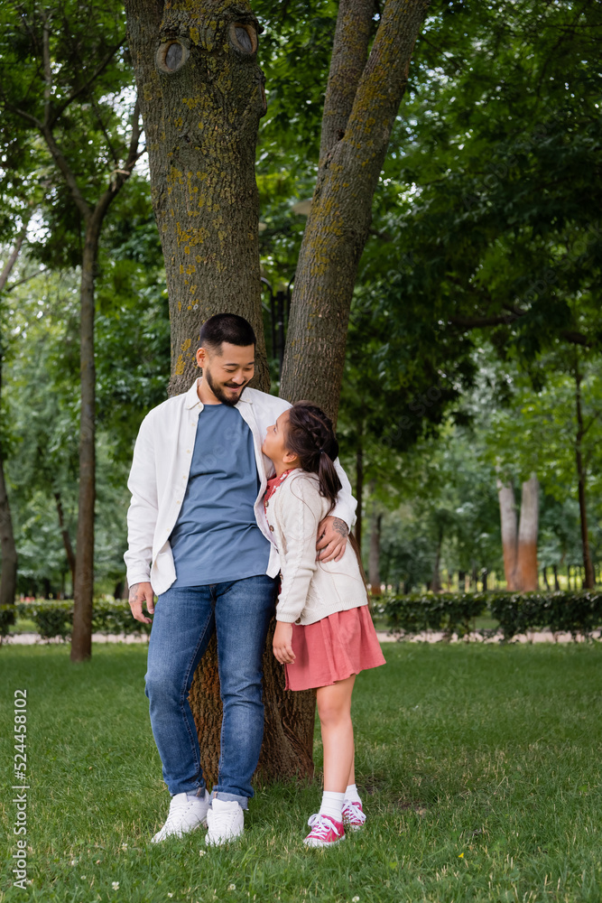 Smiling asian dad hugging and looking at daughter near tree in park.