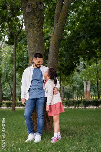 Smiling asian dad hugging and looking at daughter near tree in park. © LIGHTFIELD STUDIOS