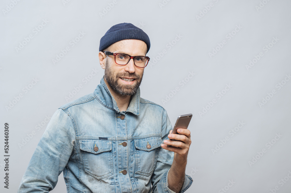 Attractive middle aged male model with bristle dressed in fashionable clothing, holds smart phone, chats with colleagues or friends in socail networks, isolated over white background. Technology Attractive middle aged male model with bristle dressed in fashionable clothing, holds smart phone, chats with colleagues or friends in socail networks, isolated over white background. Technology