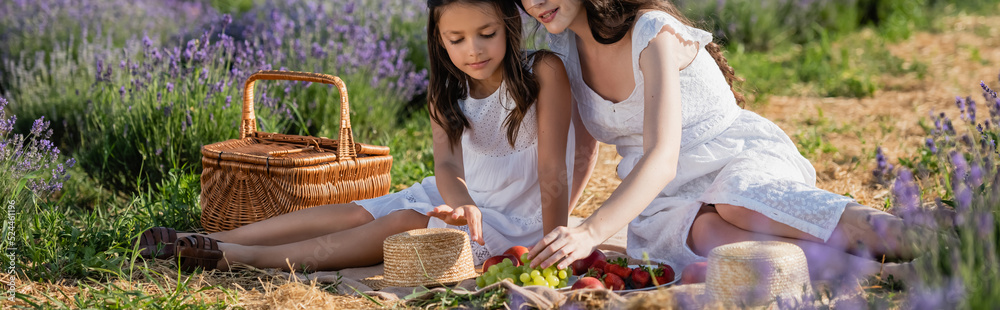 girl and mom sitting near straw basket and fresh fruits during picnic in meadow, banner.