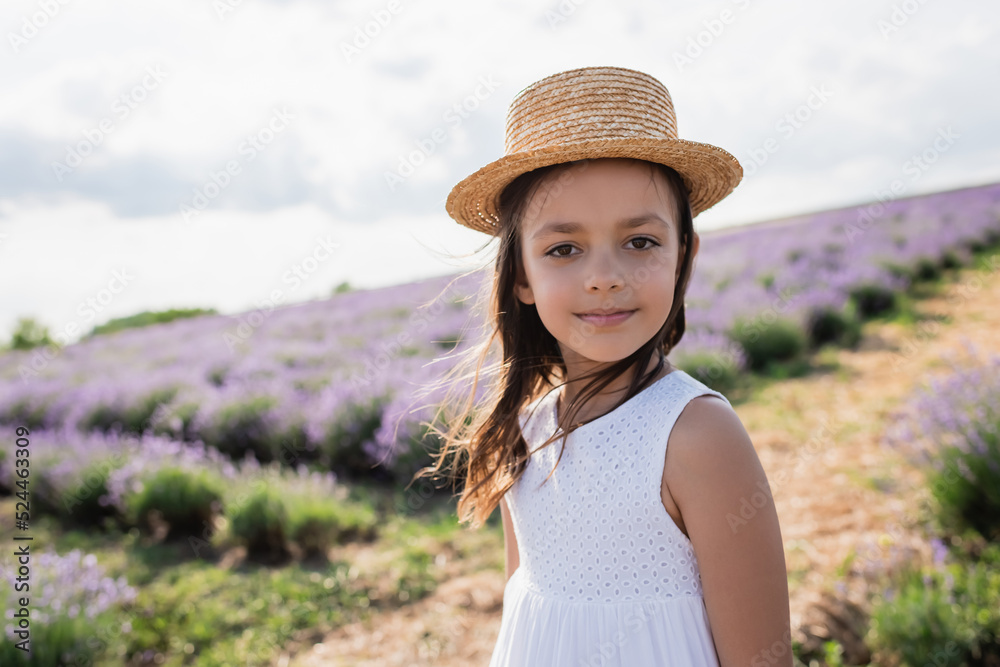 smiling girl in sun hat and white dress looking at camera in blurred lavender field.