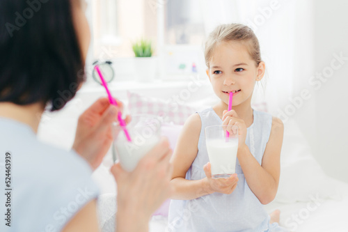 Back view of young brunette mother looks at her small daughter  drink fresh milk cocktails in bed  have delighted expressions and pleasant talk during morning  Parenthood and leisure concept