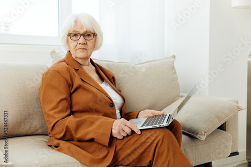 a pleasant, friendly pensioner is sitting on a beige sofa holding a laptop on her lap and smiling pleasantly while working from home in © Tatiana