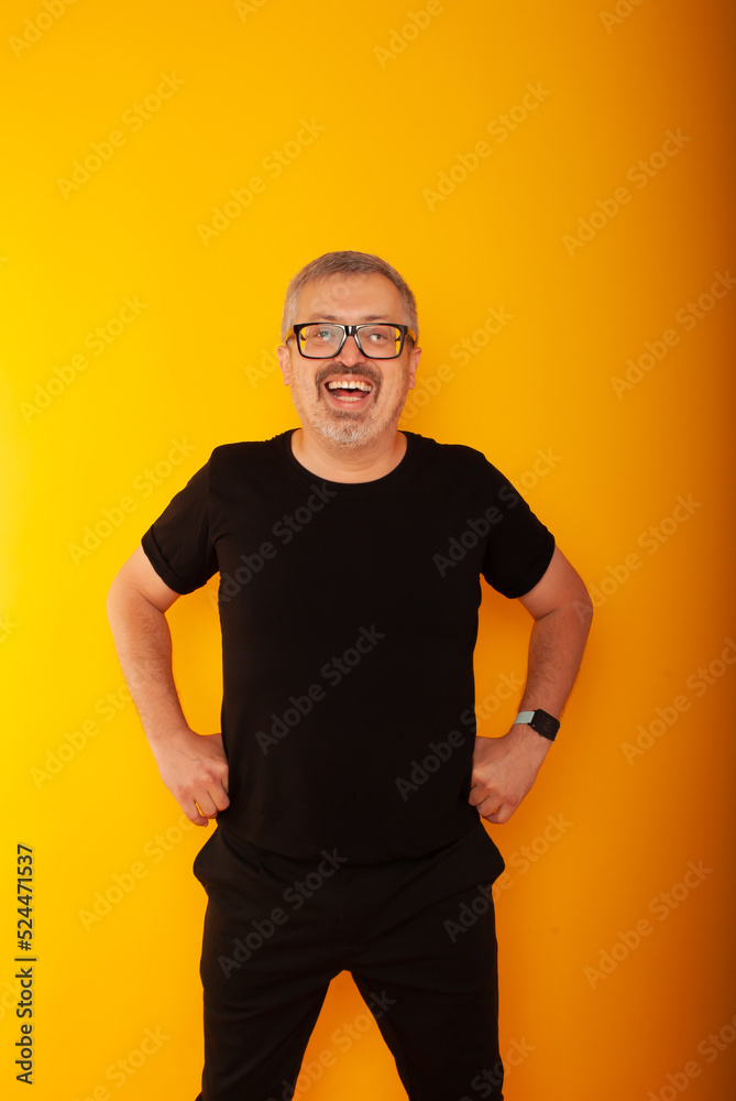 a joyful gray-haired man in black glasses and black clothes on a yellow background holds his hands on his belt a joyful gray-haired man in black glasses and black clothes on a yellow background holds his hands on his belt