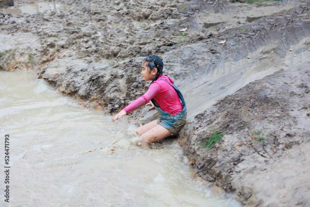 Happy dirty asian girl slider to mud puddle at outdoor summer camp learning