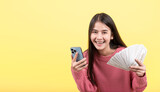 Excited beautiful woman with long brown hair standing on yellow background holding banknotes using mobile phone. Empty space.