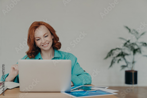 Horizontal shot of happy ginger woman freelancer works on laptop computer at home, studies online, uses software, surfs information in internet, sits at desktop, smiles positively, works remotely