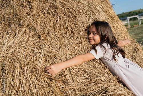 pleased brunette girl embracing haystack and looking at camera.