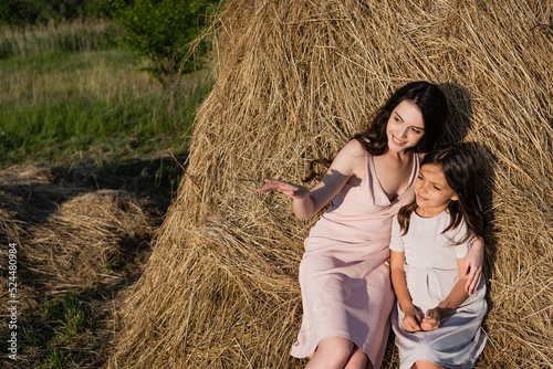 smiling woman pointing with hand while sitting on haystack with daughter.